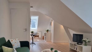 Ett mysigt vardagsrum med en grön soffa, ett soffbord och en tv på ett vitt tv-bord. Det finns en matplats med ett bord och stolar, och ett köksområde med ett vitt skåp och en växt.