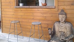 A wooden cabin with a kitchen visible through a window, two metal stools, and a Buddha statue.
