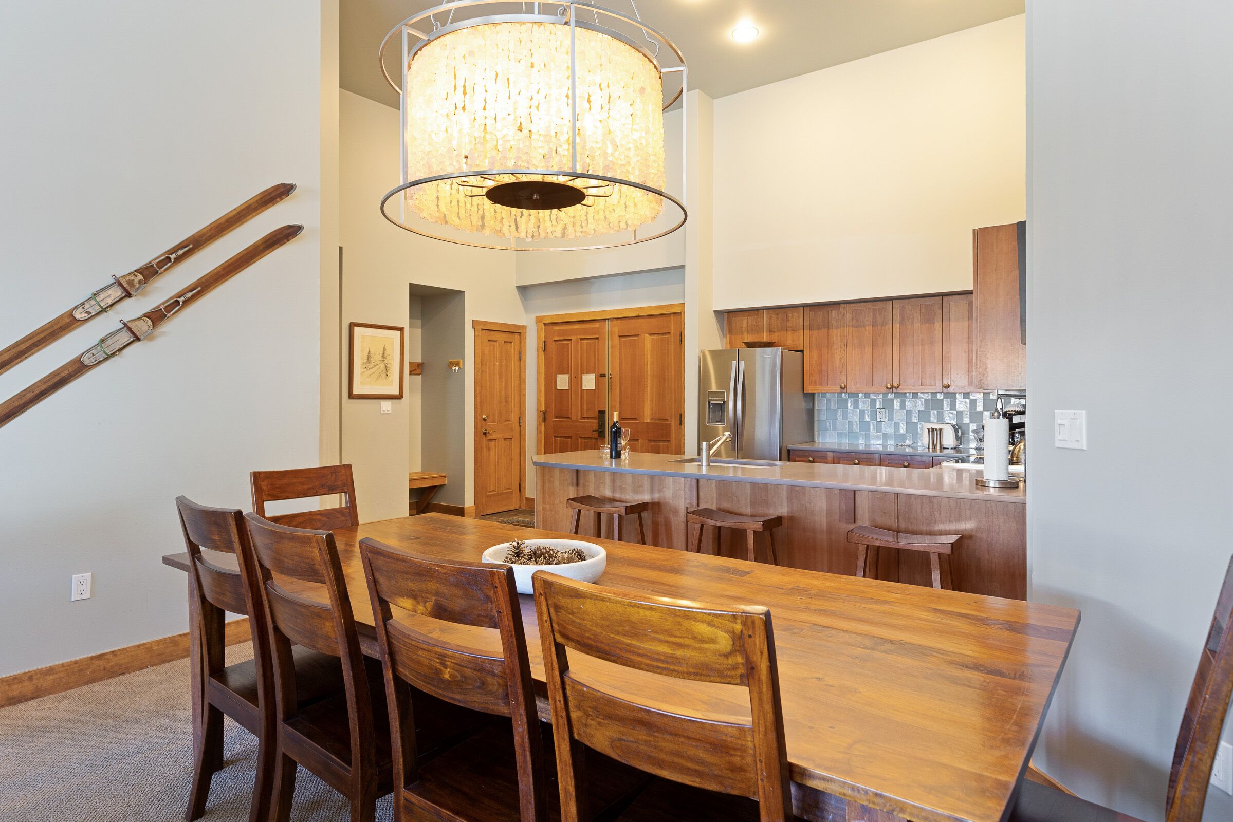 A dining area with a wooden table and chairs, a chandelier, and a kitchen in the background.