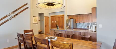 A dining area with a wooden table and chairs, a chandelier, and a kitchen in the background.