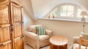 Une chambre mansardée chaleureuse, avec une armoire en bois, un canapé beige, une table ronde en bois et une fenêtre donnant sur l’extérieur.