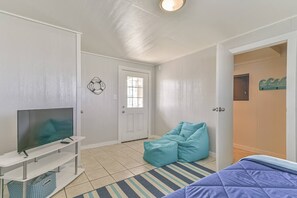 Une pièce avec un téléviseur posé sur un meuble blanc, un fauteuil en forme de poire de couleur turquoise, un tapis à rayures et un lit recouvert d’une couverture bleue.