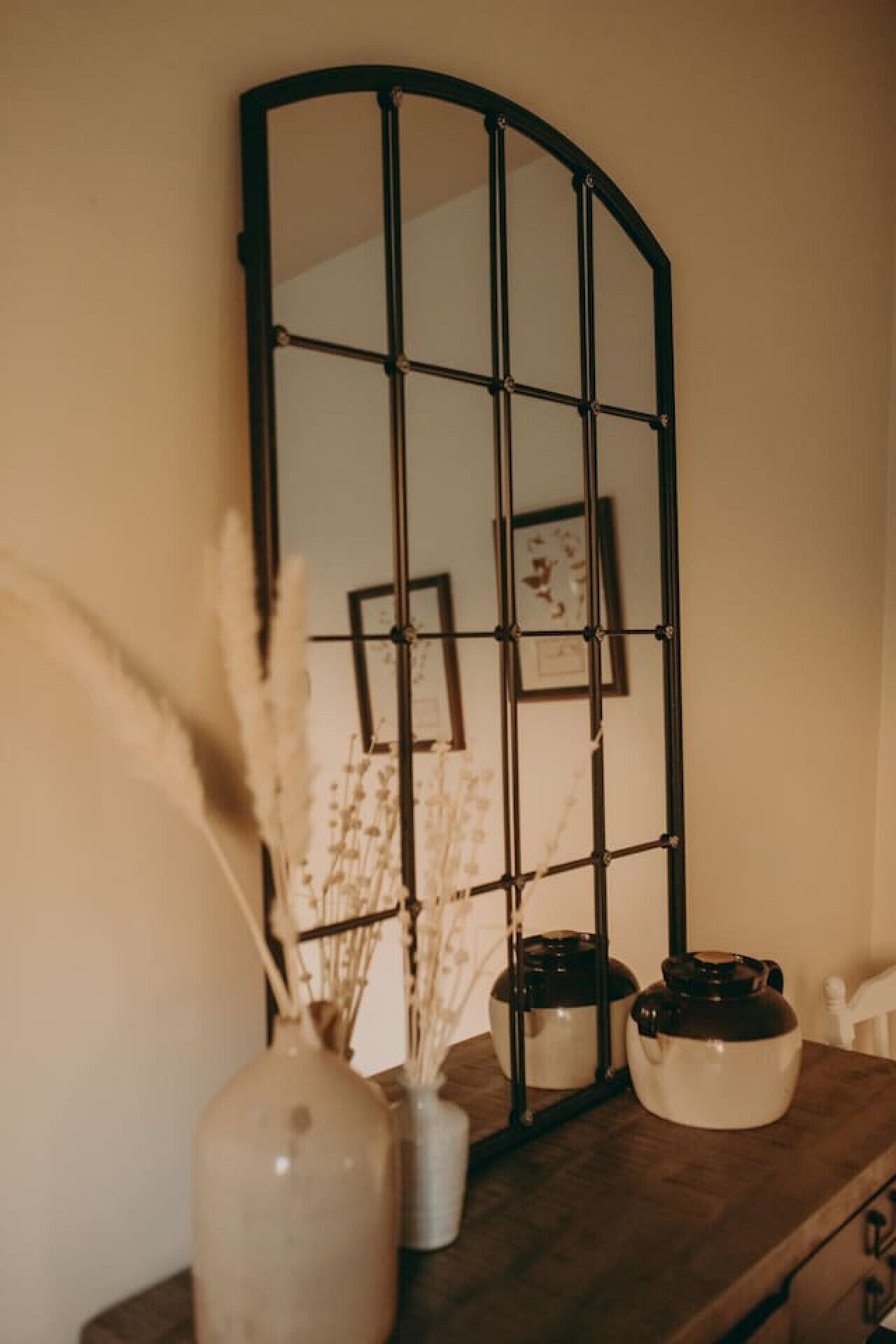 Un miroir à motifs géométriques, un vase contenant des fleurs séchées et un pot en céramique posé sur une surface en bois.