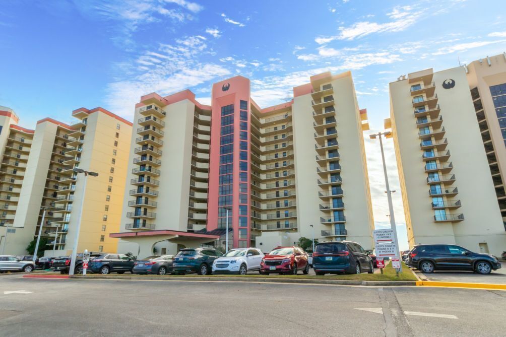 A multi-story residential building with a central red tower, parking lot, and several cars parked in front.