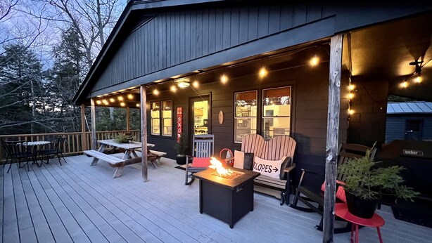 A wooden deck with picnic tables and chairs, a fire pit, and a building with a dark exterior and windows. - Cozy Manchester Getaway with Amazing Views!!! (Winhall)
