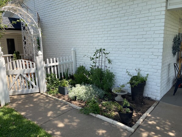 A white brick house with a white picket fence, a garden with various plants, and a small white birdbath.
