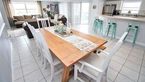 Un vaste espace de vie comprenant un coin repas avec une table en bois, des chaises blanches et un coin cuisine doté de tabourets de bar de couleur turquoise.
