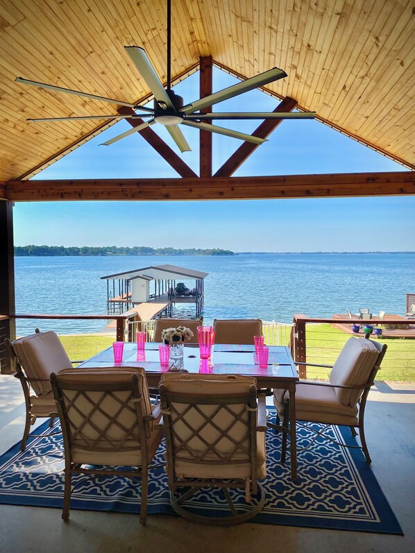 A covered outdoor dining area with a view of a lake, wooden ceiling, and a table set with chairs and pink glasses.
