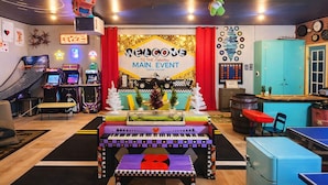 Una sala de juegos colorida con diversas máquinas recreativas, un escenario con un cartel que dice 'BIENVENIDOS AL FABULOSO MAIN EVENT', y una mesa de billar.
