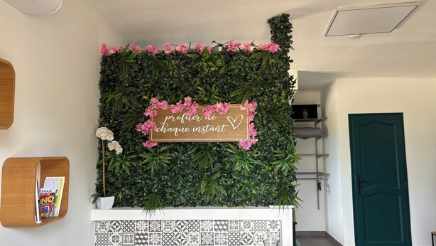 Un espace d’accueil avec un mur décoré de feuillage vert et de fleurs roses, un panneau en bois orné d’un symbole en forme de cœur et un sol carrelé.