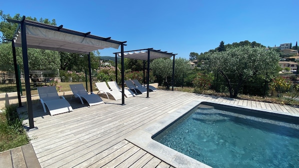 A swimming pool with a wooden deck and lounge chairs under pergolas.