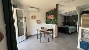 Un espace de vie compact comprenant un coin repas, un lit doté d’une tête de lit à motifs, un petit coin cuisine et un mur décoratif agrémenté de plantes et de fleurs.