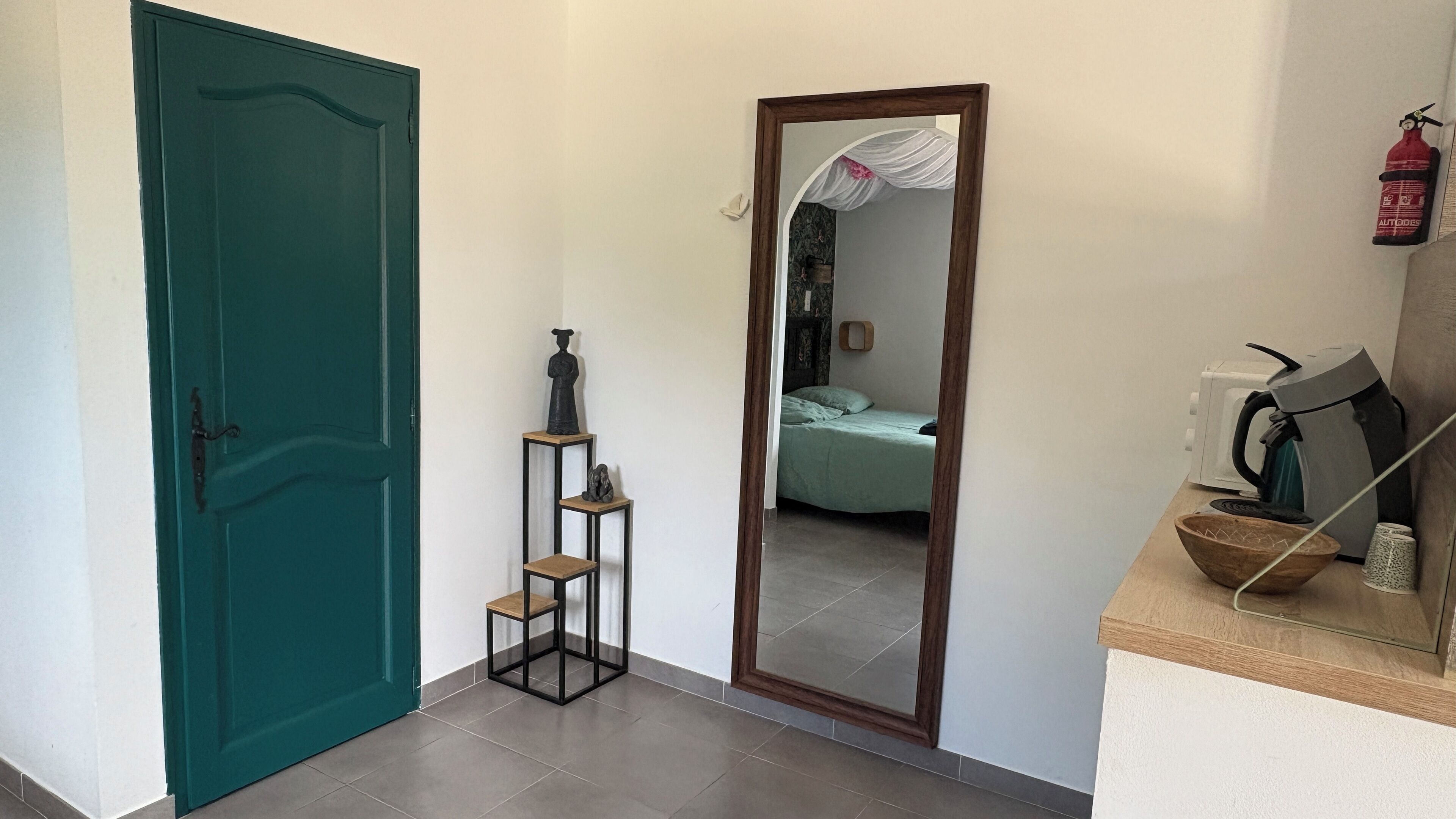 A hallway with a teal door, a mirror reflecting a bed and a room, and a kitchenette area with a kettle and cups.