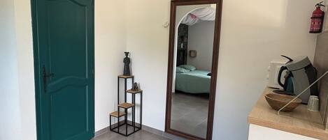 A hallway with a teal door, a mirror reflecting a bed and a room, and a kitchenette area with a kettle and cups.