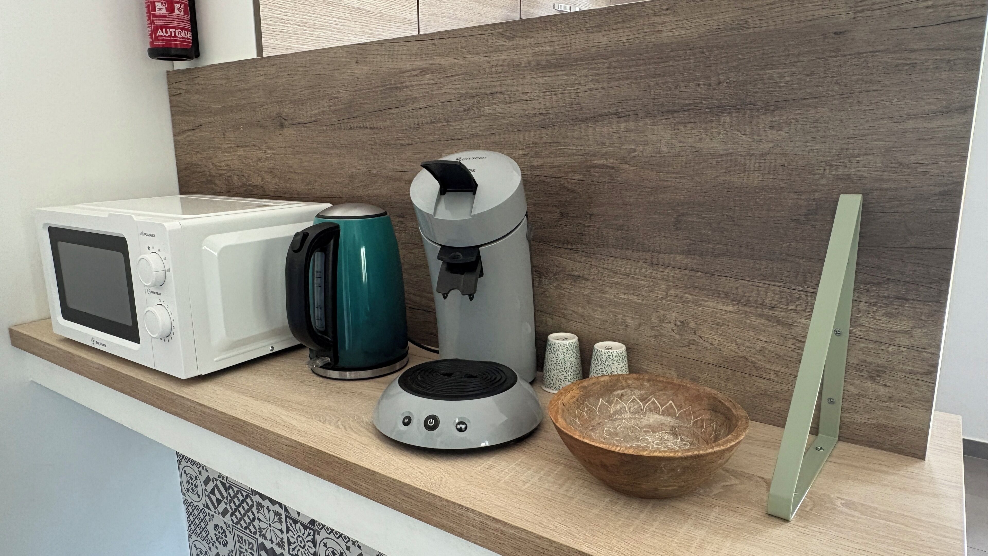 En köksbänk med en vit mikrovågsugn, en turkos elektrisk vattenkokare, en grå kaffebryggare, en liten skål och en grön skärbräda.