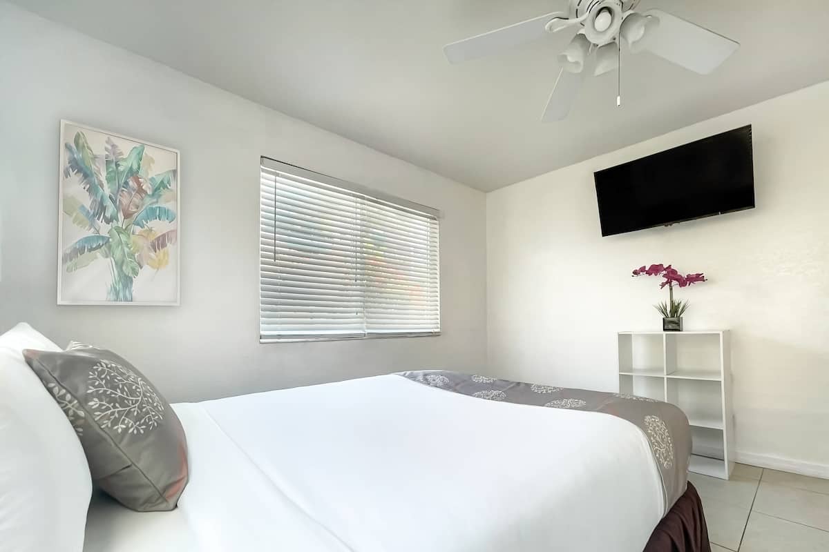 Quarto com cama, ventoinha no teto, TV fixada na parede e janela com persianas.