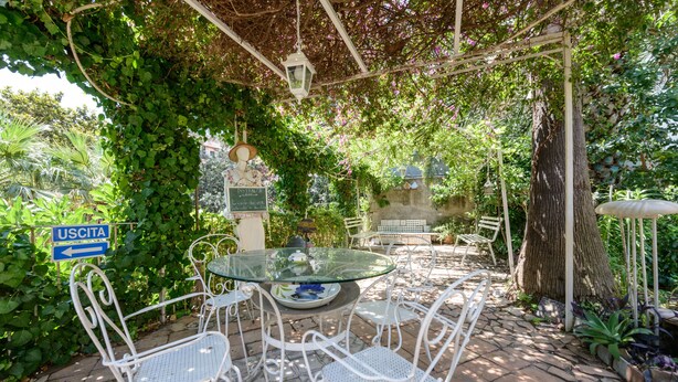 An outdoor patio with a glass table and white metal chairs, surrounded by lush greenery and a pergola. - Giardino Segreto - Via Foria 216 (Ammezzato) (Napoli)
