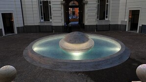 Pátio com uma fonte central, rodeado por um espaço pavimentado e defensas de pedra.