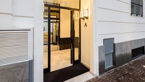 Entrada de um hotel com porta de vidro, um tapete preto e uma placa na parede com a letra 'A'.