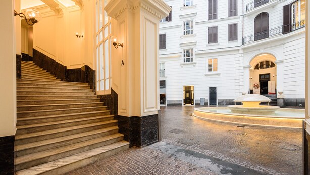 Uma escadaria imponente que leva a um andar superior, um pátio com uma fonte e a fachada de um edifício com várias janelas e sacadas.