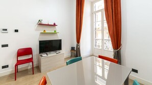 Un salon moderne avec un téléviseur à écran plat, un fauteuil rouge, une petite table et une fenêtre ornée de rideaux orange.