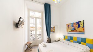 Une chambre d’hôtel équipée d’un lit, d’un bureau, d’une chaise, d’une télévision et décorée avec des œuvres d’art aux murs.