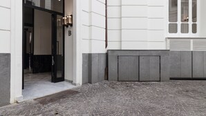 Rakennuksen sisäänkäynti, jossa on musta ovi, kirjaimella «B» varustettu kyltti ja kivilaatoitettu sisäpiha.