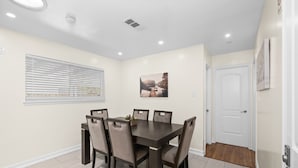 Un coin repas comprenant une table rectangulaire et six chaises, une fenêtre avec des stores, un tableau accroché au mur et une porte.