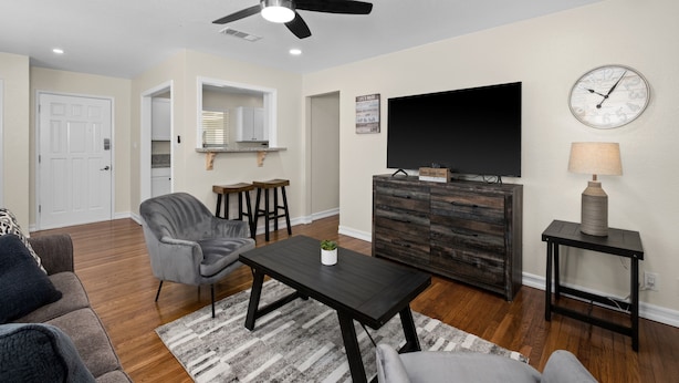 Un salon doté d’un ventilateur de plafond, d’un téléviseur à écran plat, d’une table basse en bois foncé, d’un fauteuil gris et d’une petite table d’appoint avec une lampe.