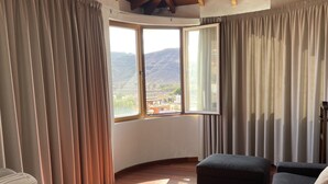 A spacious living room with wooden flooring, a large window with curtains, a sofa, and a ceiling with exposed beams. - VILLA NEILA, with heated pool  (URBANIZACIÓN PUERTO AZUL, TARAJALEJO)