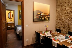 A hotel room with a bed, a bathroom, a dining area with a table set for two, and a wall-mounted picture of a street scene.