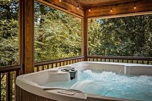 A hot tub with a wooden deck and glass walls, surrounded by lush greenery. - Dollywood Smokies Hot Tub Gameroom Fire Pit Movie (Sevierville)