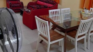 Ein Essbereich mit Glastisch und weißen Stühlen, einem roten Sofa, orangefarbenen Vorhängen und einem Deckenventilator.