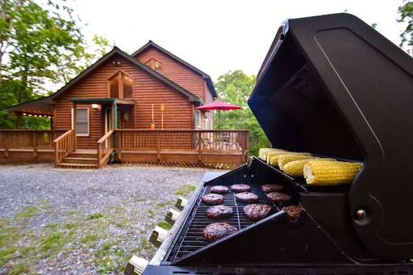 A barbecue grill with hamburgers and corn, a wooden cabin with a porch, and a gravel driveway.