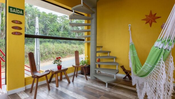 A room with a yellow wall, a spiral staircase, a hammock, and a window with a view of greenery. - Pousada Dona Elena (Penha)