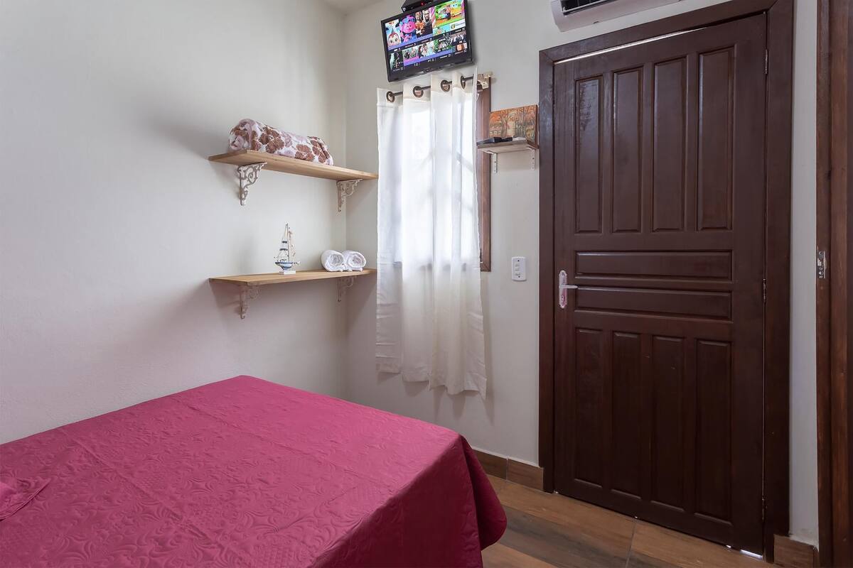 A room with a bed, a wooden door, a television mounted on the wall, and a window with curtains.