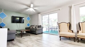 A living room with a ceiling fan, a grey sofa, a patterned armchair, a coffee table, and a TV mounted on the wall.