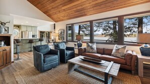 A spacious living area with a wooden ceiling, a brown leather sofa, two black armchairs, a coffee table, and large windows offering a view of trees and mountains.