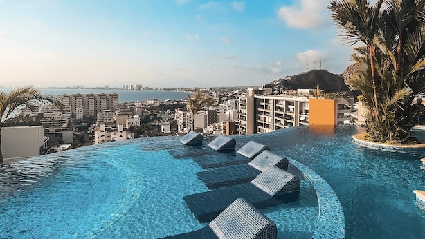 A rooftop infinity pool with lounge chairs, palm trees, and a cityscape view.