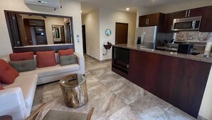 A modern living room with a sofa, a coffee table, and a TV. The kitchen has dark wooden cabinets, a microwave, and a refrigerator.