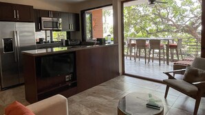 A modern kitchen with a built-in oven, microwave, and coffee maker. There is a glass-top coffee table and a sofa with a red cushion. A balcony with a table and chairs is visible through a glass door.