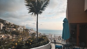 A balcony with outdoor furniture, a palm tree, and a view of the sea and buildings.