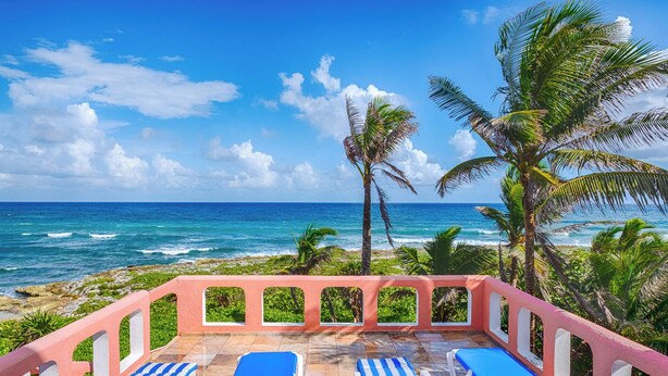 Un área de descanso en la playa con cuatro tumbonas, una barandilla rosa y vistas al océano y palmeras.