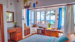 Una habitación con cama, escritorio, silla, ventana y vista al mar, además de una pequeña mesa con una planta.