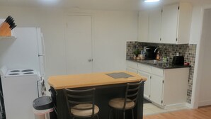 A compact kitchen with white cabinets, a small countertop, and a stove. There are two stools and a table with a beige top. A trash can is next to the table, and a patterned rug is on the floor. - Home Away Studio Basement Apartment w/Hideabed (Ogden)