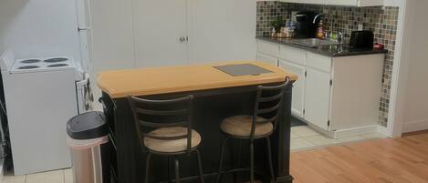 A compact kitchen with white cabinets, a small countertop, and a stove. There are two stools and a table with a beige top. A trash can is next to the table, and a patterned rug is on the floor.