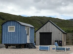 A blue wooden shed with a small window and a door, a larger shed with a black door, and a gravel ground with a wooden fence in the background. - Fishermans Hut (Harris)
