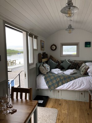 A bedroom with a bed, a window, a wooden table with glasses, and a view of the sea. - Fishermans Hut (Harris)