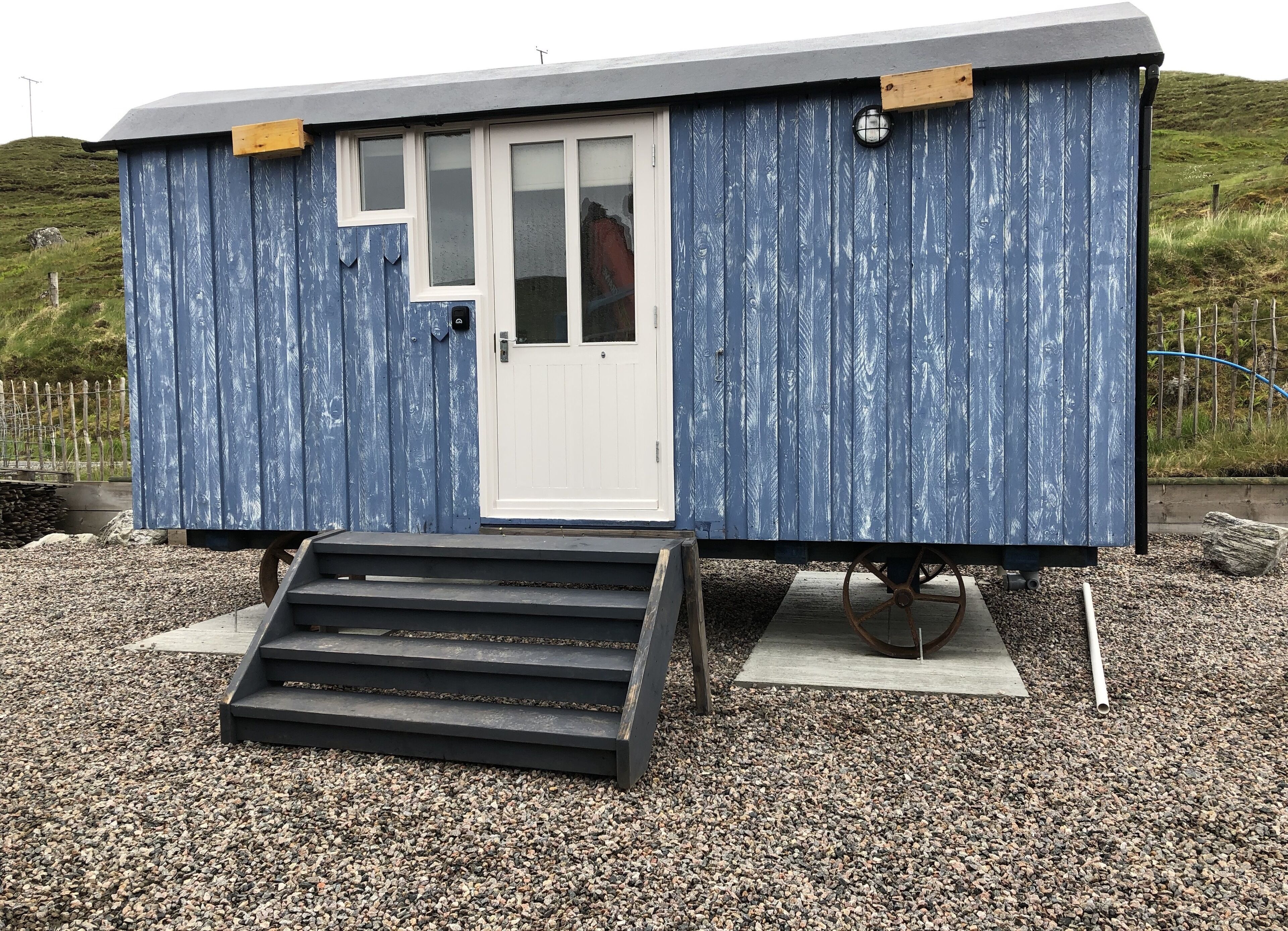 A small, single-story structure with a white door and windows, a set of steps leading to the entrance, and a gravel ground surface.