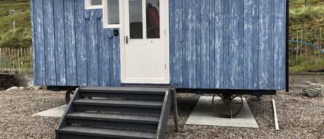 A small, single-story structure with a white door and windows, a set of steps leading to the entrance, and a gravel ground surface.